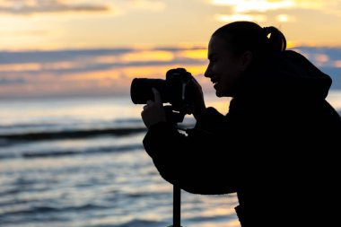 Gün batımında kamerasıyla denizi yakalayan bir kızın silueti. İlham ve özgürlükle dolu bir çerçeve.