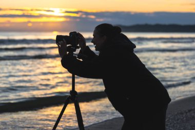 Genç bir fotoğrafçı, altın saat boyunca dalgaları yakalayan bir tripod kameraya doğru eğilir. Sıcak bir ışık ve yaratıcı günlük bir hava..