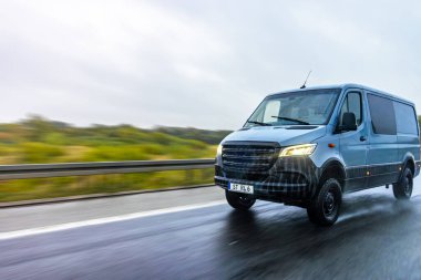 Yağmurlu bir yolda hızlı giden modern gri minibüs hareketi, özgürlüğü ve profesyonel lojistiği simgeliyor. Çekim, ıslak havada enerji ve hareketi yakalar..