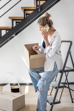 Güzel, genç bir kadın akıllı bir telefonla konuşurken elinde bir kutuyla merdivende oturuyor. Paketleri açarken birden fazla iş, sıcak gün ışığı, rahat minimalizm ve ev atmosferi.
