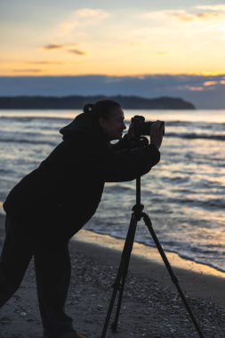 Sahilde güneş doğarken kamerayla fotoğraf çeken bir kadın, altın gökyüzüne karşı siluet sakin ve yaratıcılığını gösteriyor..