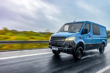 Yağmurlu bir otoyol boyunca mavi bir ticari minibüs hızlanır. Geniş perspektif, parlak farlar ve su yolları dinamik taşımacılığı ve yola hazır olmayı vurguluyor.