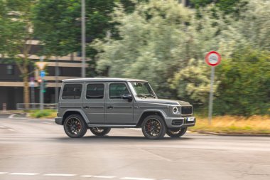 Arka planda yeşil ağaçlar ve modern binalar bulunan gri bir SUV, lüks, güç ve konforu temsil ediyor..