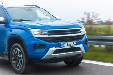 Islak bir otoyolda giden mavi bir kamyonetin yakın görüntüsü. Yağmur damlaları ve hareket bulanıklığı gücü, yol tutuşunu ve güvenilir hareketliliği vurgular.