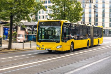 Şehir merkezinde uzun ve eklemli sarı bir otobüs hareket bulanıklığı ile hızla ilerliyor, verimli ve sürdürülebilir kamu hareketliliğini simgeliyor.