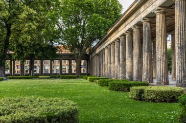 Yeşillik ağaçlar ve manikürlü çimenler tarafından çerçevelenmiş klasik bir taş sütun, antik mimariyle doğayı birleştirir. Yumuşak gün ışığı sütunların dokusunu güçlendirir ve sakin, zarif bir atmosfer yaratır..