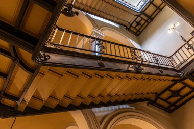 Aşağıdan çekilen bir mimari çekimde süslü metal korkuluklar ve sıcak ışıklarla donatılmış, zamansız zarafet ve derinliği çağrıştıran klasik bir merdiven görülüyor..