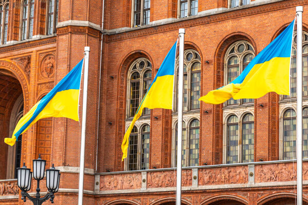 Три украинских синих и желтых флага - волны у богатого кирпичного здания, символизирующие национальную гордость, стойкость и единство.