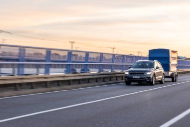 Karanlık bir SUV, kapalı bir römorku yumuşak günbatımı renklerine ve uzak ufuk çizgisine sahip bir viyadük üzerinde taşıyor. Hareket kabiliyeti, küçük lojistik ve şehir içinde seyahat.