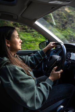 Yeşil kapüşonlu bir kadın dağ yolunda modern bir araba kullanıyor. Doğal ışık ve bereketli yeşillik bir sakinlik, özgürlük ve doğa ile bağlantı hissi yaratır..