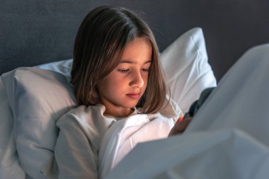 Küçük kız beyaz bir yatakta uzanıyor ve telefon ekranına odaklanıyor. Yumuşak akşam ışığı, güvenlik hissi ve acele etmeden göz atma hissi..