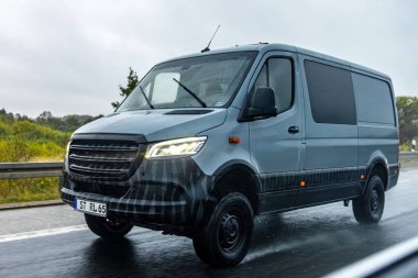 Yağmurda ıslak bir otoyolda gri bir minibüs sürüyor. LED farlar, tekerlek spreyi ve hareket bulanıklığı lojistik, güvenilirlik ve yoldaki hizmeti vurguluyor.