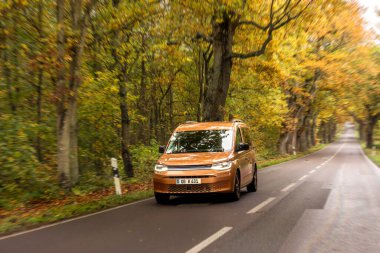 Minibüs, güz ağaçlarının altında düz bir kırsal otoyol boyunca gidiyor. Uygulamayla şehir dışı yolculuğunun rahat havasını birleştiriyor..