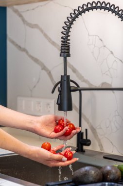 Modern bir mutfakta, bir kadın bir avuç dolusu kiraz domatesi akan suyun altında duruluyor. Parlak gün ışığı ve damlacıklar yemek pişirmeden önce tazeliği ve hijyeni vurgular..