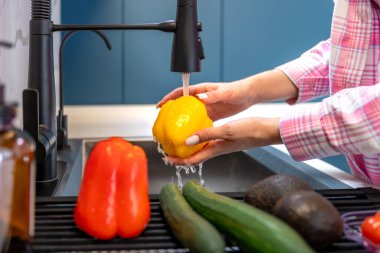 Modern bir mutfakta, bir kadın yanında başka ürünlerle birlikte akan suyun altında sarı dolmalık biberi duruluyor. Sahnede hijyen hazırlık ve sağlıklı yemekler için taze malzemeler vurgulanıyor..