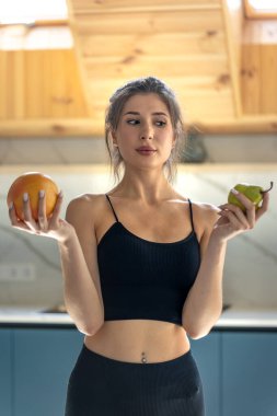 Sportif bir kadın her iki elinde meyvelerle mutfakta duruyor ve yemek planını düşünüyor. Yumuşak gün ışığı ve soluk renkler dengeyi ve temiz yiyeceği sağlar..