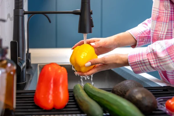 Modern bir mutfakta, bir kadın yanında başka ürünlerle birlikte akan suyun altında sarı dolmalık biberi duruluyor. Sahnede hijyen hazırlık ve sağlıklı yemekler için taze malzemeler vurgulanıyor..