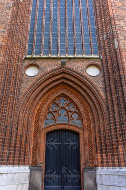 Üsteki uzun gotik pencereli ve karanlık kapılı kırmızı tuğlalı duvar dikey görünüyor. Tekrarlayan çizgiler ve zengin dokular kilise cephesine güçlü bir mimari ritim ve şehir havası veriyor..