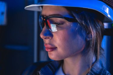 Kameraya yansıyan ekran verileri ile güvenlik gözlüğü takan bir kadın elektrikçinin yakın çekimi. Yumuşak mavi ışıklandırma ai siber güvenlik ve dijital izleme temaları için odaklanmış bir ortam oluşturur.