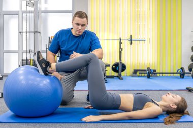 Doktor teknik ve leğen kemiğini incelerken bir hasta, ayakları sabit bir topun üzerinde mavi bir mindere uzanıyor. Temel eğitim, denge ve akıllı iyileşme için modern rehabilitasyon sahnesi.