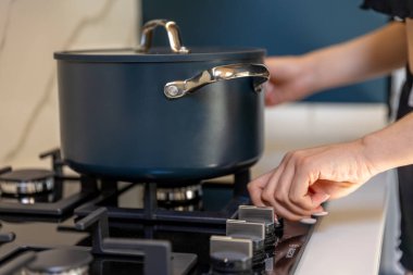 A close view shows a hand rotating a gas stove control beside a dark lidded pot. Crisp detail signals the start of cooking and a warm kitchen mood.