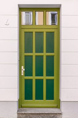 Zeytin çerçeveli ince yeşil bir kapı ve panellerde yakınlardaki binaları yansıtan küçük cam camlar bulunuyor. Temiz beyaz cephe, minimalist şehir tarzını ve konut hissini arttırır..