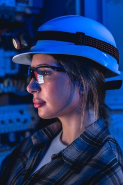 Dağıtım ekipmanlarının yanında, far ve güvenlik gözlüğü takan kadın teknisyen profili. Koyu mavi ışık, modern şebekeler için gece vardiyası güvenilirliğini ve akıllı izlemeyi çağrıştırıyor.