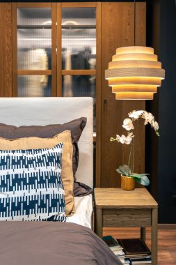 Yumuşak yastıklar ve sıcak ışık altında bir orkide ile yaşamaya hazır olmak samimi bir his yaratır. Ahşap gardıroplar ve karanlık duvarlar derinlik ve kaliteli atmosfer ekler..