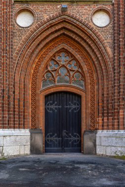 Koyu ahşap kapıları ve desenli pencereleri olan uzun gotik bir kapı kırmızı tuğla duvarlara dayanır. Karmaşık dekorlar ve ağır şekiller kilisenin girişine güçlü bir tarihi hava katıyor..