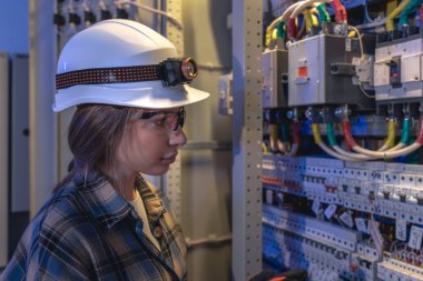 Kasklı, farlı bir kadın elektrikçi, havalı mavi ışığın altında santrali dikkatle kontrol ediyor. Sahne, sanayi altyapısının güvenliği, güvenilirliği ve modernizasyonunu vurguluyor.