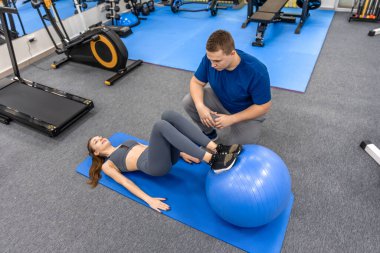 Doktor teknik ve tempoyu izlerken hasta dengeli bir topla bacak egzersizi yapıyor. Geniş rehabilitasyon salonu güvenlik işlevsel iyileşme ve bakımı vurguluyor.