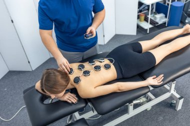 Muayene sırasında eldivenli bir doktor omuz ve köprücük kemiği bölgesini incelerken kadın hasta hislerine odaklanıyor. Nötr klinik içi ağrı kesici ve iyileşme konusunda tıbbi içeriğe uygun.