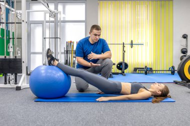 Doktor güvenli hareket ve formda koçluk yaparken hasta ayakları dengeli bir topun üzerinde yatıyor. Parlak rehabilitasyon salonu atmosferi, çekirdek denge, denge, refah ve uzun yaşam odaklı.