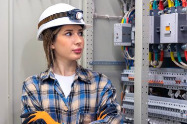 Kasketli bir kadın mühendis açık bir telefon santralinin yanında duruyor ve içeride organize kablolar var. Görüntü düzen, kalite kontrol ve kadınların sanayiye olan güvenini aktarıyor..