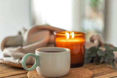 Mantar altlığı üzerinde seramik bardak yanan bir mum ve ahşap bir masada yumuşak örgü battaniyesi. Sıcak gün ışığı ve sakin tasarım sağlığa uygun yavaş yaşayan ve dikkatli ev içeriği.