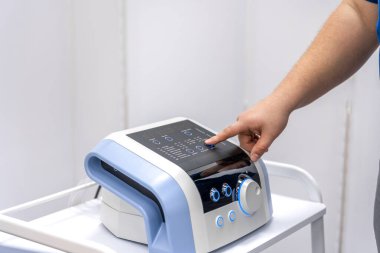 Rehabilitasyon merkezindeki modern bir pressoterapi ünitesini kapatın. Doktor eli kontrol panelinden bir program seçer. Temiz beyaz arka plan, hassasiyet hissi, sağlık teknolojisi eğilimi.