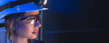 Sert şapka takan ve koruyucu gözlük takan bir kadın elektrik panelleriyle teknik odaya odaklanmış. Dramatik mavi ışık profesyonelliği vurguluyor ve güvenlik ve enerji endüstrisi kavramlarını destekliyor.