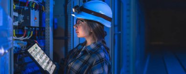 One woman in hard hat and gloves analyzing data on tablet near electrical switchboard. Blue atmospheric lighting creates modern high tech mood for energy and digital control concepts.