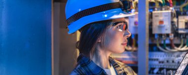 One woman in hard hat and protective glasses reviewing tablet near electrical panel. Dramatic blue lighting highlights high tech atmosphere for safety, energy and industrial innovation concepts.