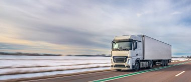 Gündüz vakti karlı kırsal araziyle çevrili kış otoyolu boyunca mal taşıyan modern beyaz yük kamyonu. Lojistik, kargo taşıma ve teslimat kavramı.