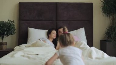 Mutlu bir genç Aile büyük bir yatakta yatarken ve güzel sohbet; küçük bir kız için çalışan gelir ve yatağın üzerine atlar, Anne Çocuk kucakla.