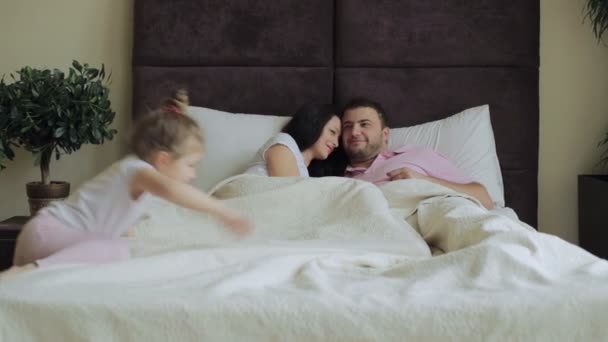 Een gelukkig jong gezin is liggend op een groot bed en chatten mooi; een meisje komt lopen naar hen en springt op het bed, de ouders knuffel het kind. — Stockvideo