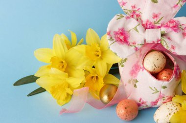 Dekoratif sepette renkli Paskalya yumurtaları ve mavi arka planda bir buket sarı narsist.
