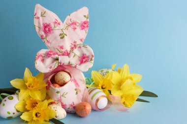 Dekoratif sepette renkli Paskalya yumurtaları ve mavi arka planda bir buket sarı narsist.