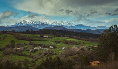 Kuzey İspanya 'daki Cantabrian kırsalının panoramik manzarası, yuvarlanan yeşil tepeler, dağınık kırsal evler, ormanlar ve karla kaplı dağlar. Barışçıl tarım manzarası doğayı gösteriyor, geleneksel köy mimarisini.
