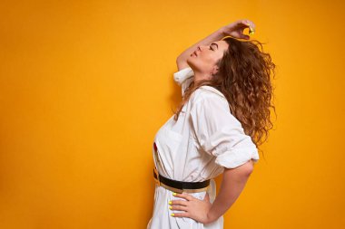 Genç kıvırcık saçlı, saçları dolu bir kız sarı bir arka planda dikiliyor ve yukarı bakıyor. Bir eli kemerinde, diğeri de saçlarını düzleştiriyor..