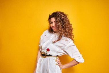 Genç, seksi, kendine güvenen, kıvırcık saçlı, beyaz gömlekli, sarı arka planda poz veren bir kız. Bir eli onun tarafında duruyor. Sarı arka planda genç bir kızın portresi.