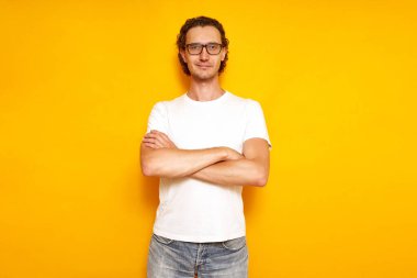 Genç, sakin, ciddi bir adam, gözlüklü, küçük bir gülümseme, beyaz tişörtlü, mavi kotlu, kolları göğsünde, izole edilmiş sarı bir arka planda duruyor. kopyalama alanı. insan kavramı