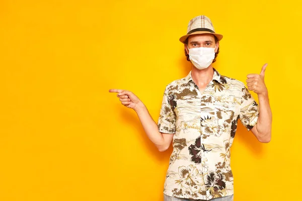 Male vacationing tourist in medical mask points his finger at an empty area and stands with his thumb raised up isolated on yellow background dressed in shirt, hat. concept rest, vacation, travel — Stock Photo, Image