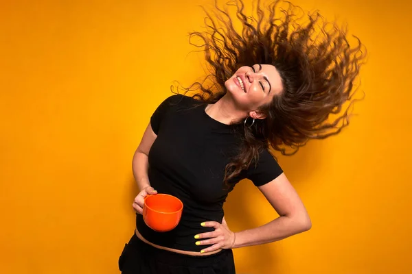 Young woman in casual clothes with an orange mug in her hand is happy with coffee she has drunk and enjoys cheerfulness in morning, throwing her hair back. stands isolated on yellow background — Stock Photo, Image